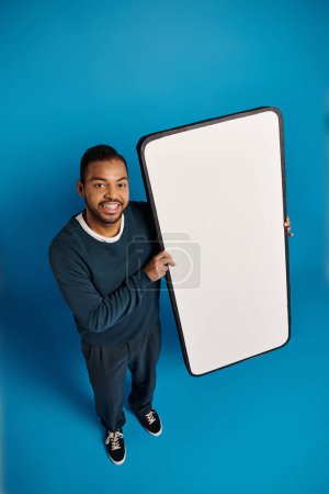 Photo for Top shot of african american young man holding white banner or smartphone in hands against blue - Royalty Free Image
