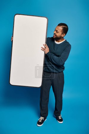 smiling african american man holding and looking at smartphone mockup on blue background