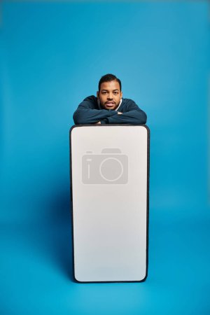 handsome african american young man leaning on smartphone mockup with folded arms on blue background