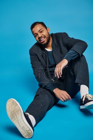 fröhlicher afrikanisch-amerikanischer Mann sitzt mit gebeugtem Bein und blickt in die Kamera auf blauem Hintergrund