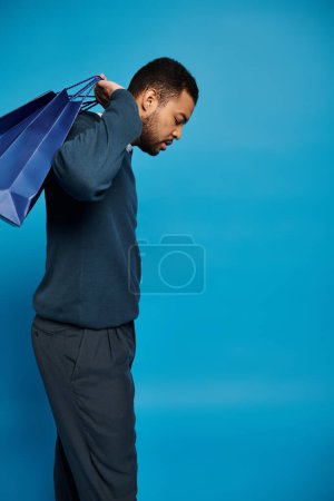Afrikanisch-amerikanischer Mann im dunkelblauen Outfit schaut nach unten und hält Einkaufstaschen über der Schulter