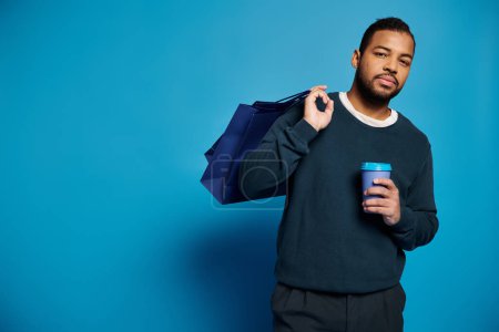 attractive african american man holding paper cup with shopping bag over shoulder on blue background