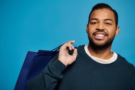 Porträt eines fröhlichen afrikanisch-amerikanischen jungen Mannes mit Einkaufstasche über der Schulter auf blauem Hintergrund