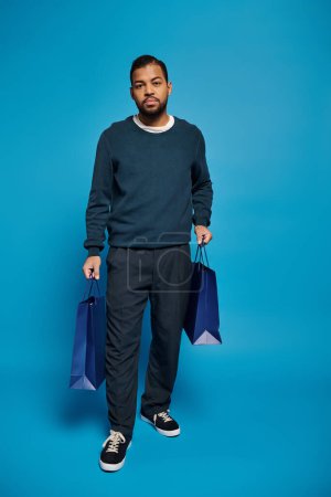 guapo afroamericano hombre caminando con bolsa de compras en las manos sobre fondo azul