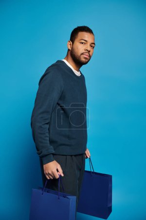 charming african american man in 20s standing sideways with shopping bag in hands on blue background
