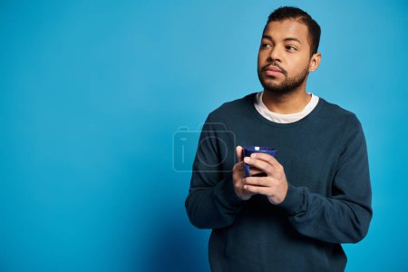 Photo for Thoughtful african american man in 20s holding cup in hands and looking to side on blue background - Royalty Free Image