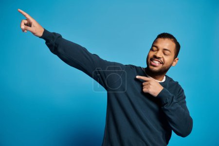 Photo for Cheerful african american man in his 20s showing fingers to side with closed eyes on blue background - Royalty Free Image