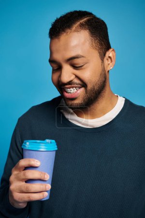 Porträt eines lächelnden afrikanisch-amerikanischen jungen Mannes, der auf einen Pappbecher in der Hand auf blauem Hintergrund blickt
