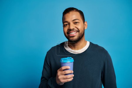 Téléchargez les photos : Portrait de charmant jeune homme afro-américain regardant à la caméra avec tasse en papier à la main - en image libre de droit