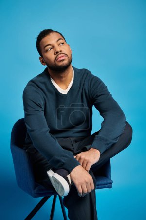 thoughtful african american man in his 20s sitting on chair and leaning to legs on blue background