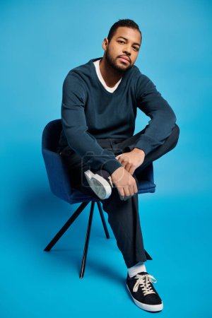 charmant homme afro-américain dans la vingtaine assis sur une chaise et penché à la jambe pliée sur fond bleu