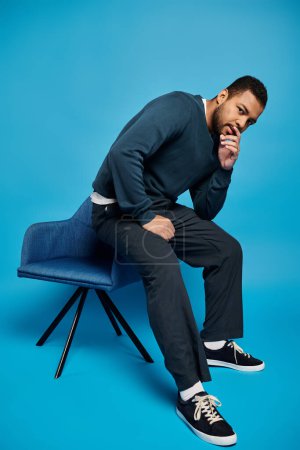 attractive african american man sitting on back of chair and touching to face on blue background