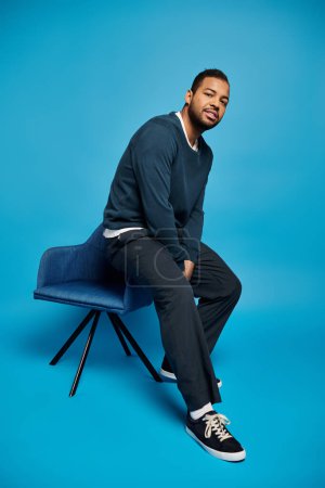 atractivo afroamericano hombre en traje oscuro relajado sentado en el respaldo de la silla sobre fondo azul