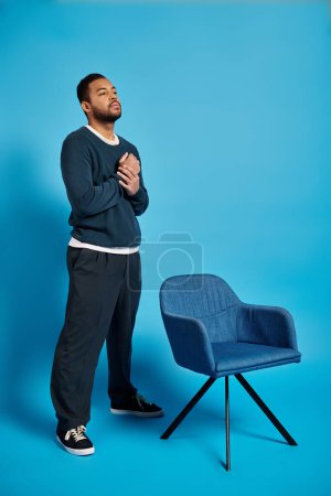 encantador hombre afroamericano con las manos en el pecho de pie cerca de la silla sobre fondo azul