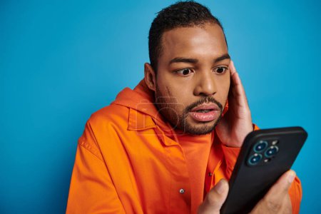 Téléchargez les photos : Astonished african american man scrolling social media in smartphone on blue background - en image libre de droit