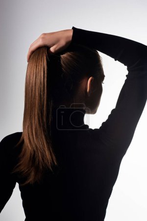 A young beautiful woman with long hair holds her hair while wearing a black turtleneck.