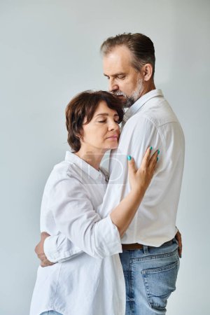Foto de Atractiva mujer de mediana edad abrazando al hombre maduro en camisa blanca con amor sobre fondo gris - Imagen libre de derechos