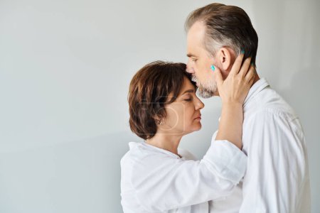 Handsome middle aged man kissing forehead of brunette woman with love on grey background