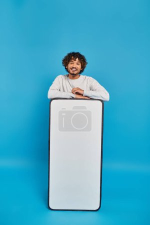 Téléchargez les photos : Un homme se tient caché derrière un gros objet blanc dans un décor studio sur fond bleu. - en image libre de droit