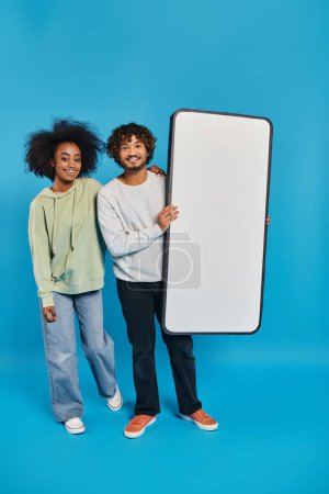 Téléchargez les photos : Un couple diversifié d'étudiants se tiennent côte à côte près d'une maquette de smartphone dans un studio, mettant en valeur la diversité culturelle sur un fond bleu. - en image libre de droit