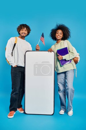 A pair of diverse individuals, multicultural students, stand near a smartphone mockup in a studio against a blue background.