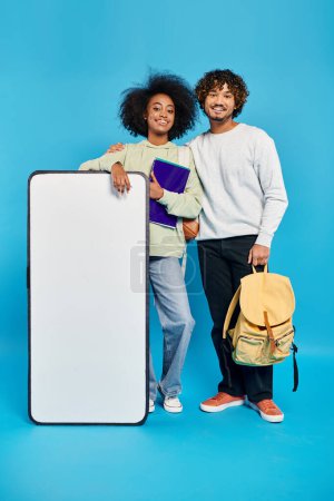 Una pareja diversa de estudiantes de pie junto a una maqueta de teléfonos inteligentes en un estudio con un fondo azul.