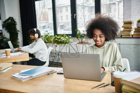 A woman of diverse background focused on her laptop in a modern coworking space among other multicultural students.