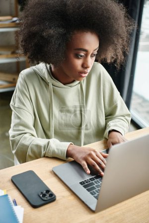 A black woman sits at a table, intensely focused on her laptop in a modern coworking space