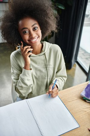 Foto de Una mujer negra absorta en una llamada telefónica mientras está sentada en una mesa adentro. - Imagen libre de derechos