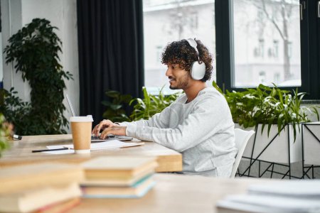 hombre indio sentado en una mesa, absorto en su portátil, con auriculares, inmerso en su trabajo en un moderno espacio de coworking.