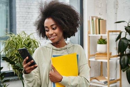 Una mujer de varias etnias sostiene una carpeta mientras envía mensajes de texto en un teléfono inteligente en un espacio de coworking moderno.