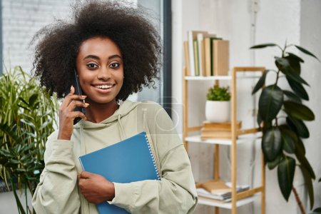 Eine schwarze Frau unterschiedlicher ethnischer Zugehörigkeit spricht auf einem Handy, während sie einen Ordner in einem modernen Coworking Space hält.