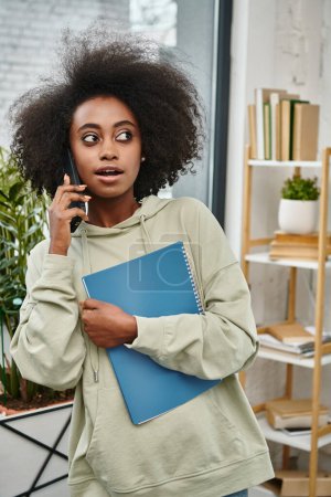 Schwarze Frau mit unterschiedlichem Hintergrund spricht auf einem Handy, während sie einen Ordner in einem modernen Coworking Space hält.
