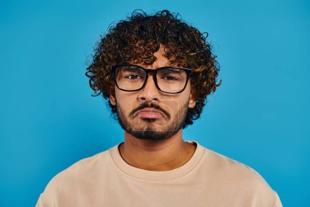 Un estudiante indio con el pelo rizado y gafas posa con confianza sobre un telón de fondo azul en un entorno de estudio.