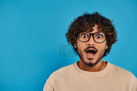 Eine indische Studentin mit lockigem Haar und Brille sieht vor blauem Hintergrund überrascht aus.