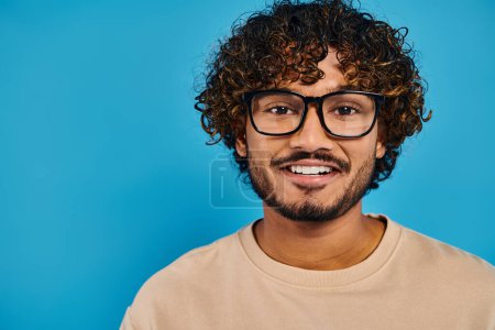 Ein gelehrter indischer Student mit lockigem Haar und Brille steht selbstbewusst vor einem leuchtend blauen Hintergrund..