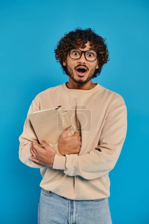 shocked Indian student with curly hair and glasses, standing in casual attire, holding a folder against a blue backdrop in a studio.