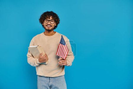 Un homme tient fièrement un livre et un drapeau américain, debout sur un fond bleu dans un studio.