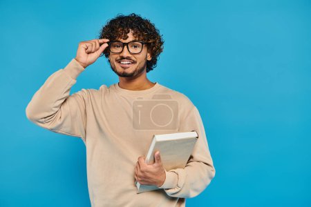 Téléchargez les photos : Un étudiant indien en tenue décontractée, portant des lunettes, tenant une tablette dans un studio avec une toile de fond bleue. - en image libre de droit
