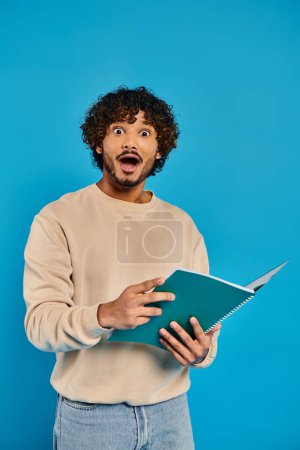 Photo for An Indian student with curly hair holding a book, standing casually against a blue backdrop in a studio. - Royalty Free Image