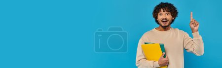 Photo for An Indian student stands in casual attire, holding a book and pointing upwards against a blue backdrop. - Royalty Free Image