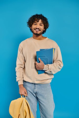A young Indian man with curly hair holding a book, standing against a blue backdrop in a studio.