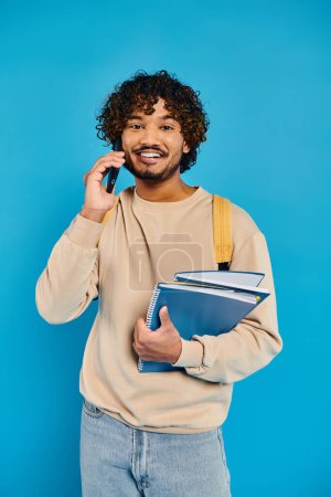 A man, dressed casually, holds a book while talking on his cell phone against a blue backdrop.