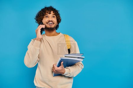 An Indian student in casual attire stands against a blue backdrop, holding a folder and talking on a cell phone.
