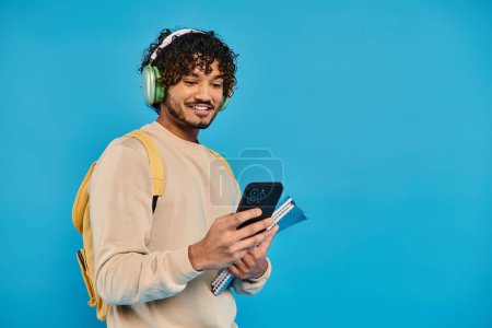 Photo for An Indian student standing, wearing headphones, and holding a book against a blue backdrop in a studio setting. - Royalty Free Image
