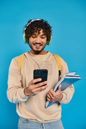 Ein indischer Student in Freizeitkleidung hört Musik über Kopfhörer, während er sein Handy vor blauem Hintergrund hält.