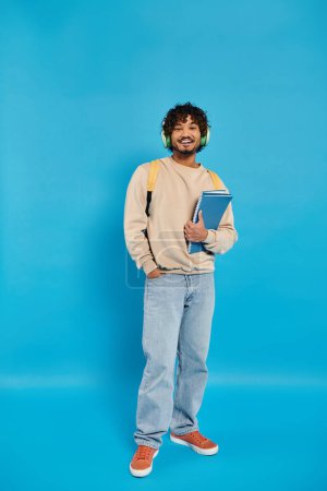An Indian student standing in casual attire, holding a book in his hands against a blue backdrop in a studio.