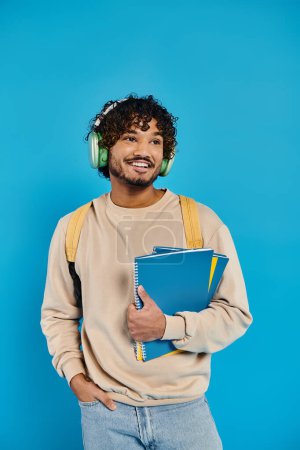Ein indischer Student steht vor blauem Hintergrund, trägt Kopfhörer und hält ein Buch in der Hand, eine harmonische Mischung aus Musik und Literatur..