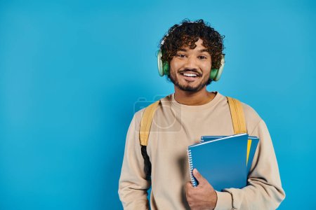 heureux homme indien portant des écouteurs, tenant des livres et souriant sur fond bleu