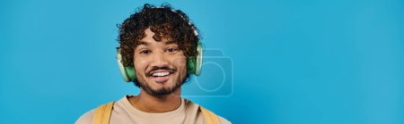 Téléchargez les photos : Heureux homme indien portant des écouteurs et souriant sur fond bleu - en image libre de droit